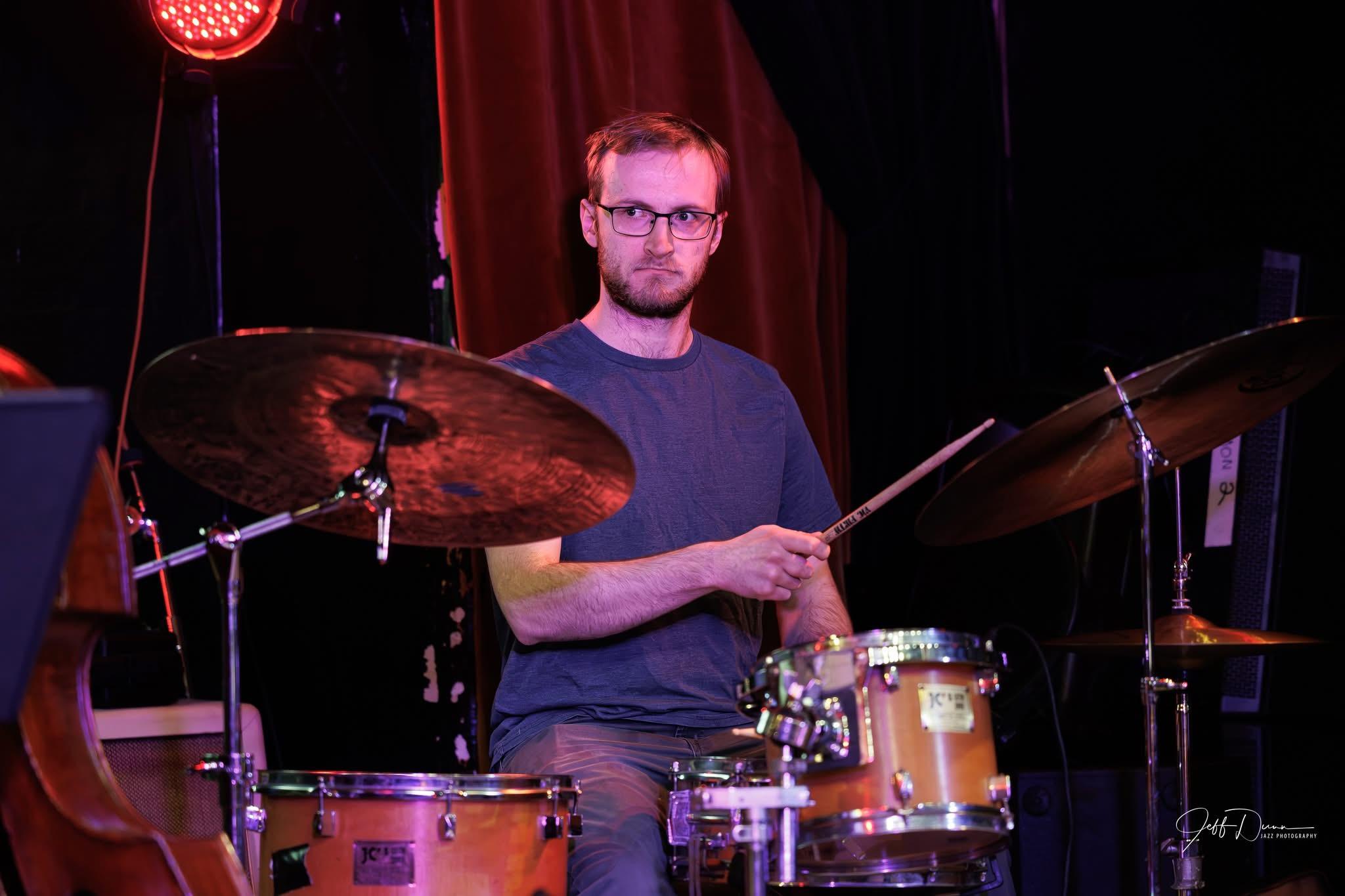 Spencer Perilloux seated at a drum set in a theatre setting.